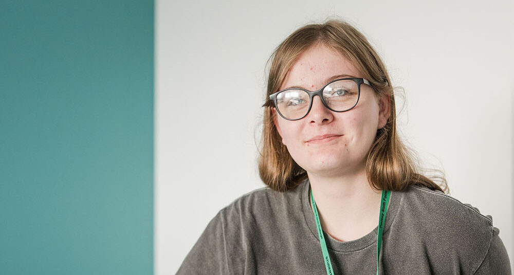 Student wearing grey top and glass in classroom with laptop smiling