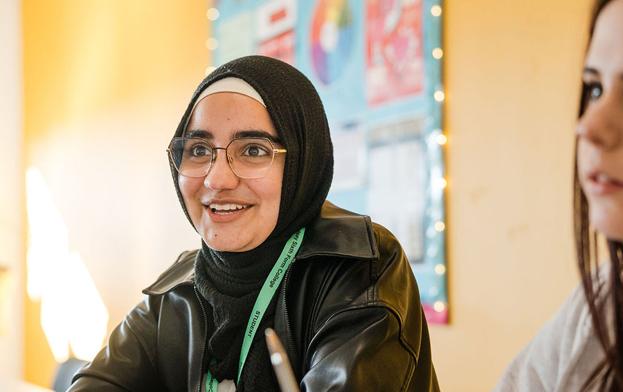 Student wearing hijab headscarf sitting down smiling with other students at the background in the classroom