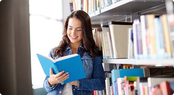 Student stood next to shelves of books in library, reading a book with a blue cover