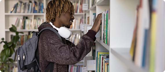 Student in library wearing rucksack and headphones looking through books on a shelf