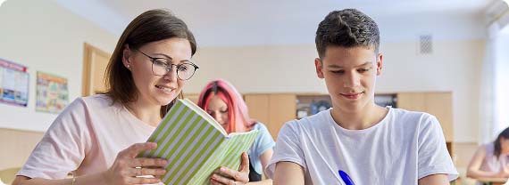 Teacher and students sat in classroom at desks reading from a book and making notes with a pen