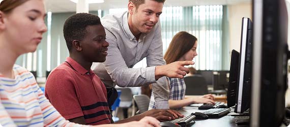 Students sat in computer room with teacher leaning over and explaining something on one of the computer screens