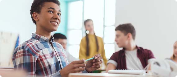Student sat at a desk in a classroom holding a pen whilst looking up and smiling