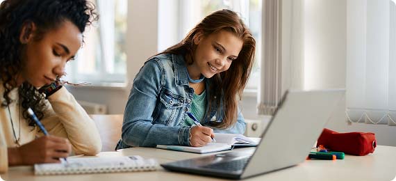 Two students sat at a desk in a classroom writing notes whilst sharing a laptop screen
