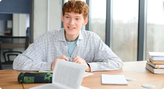 Student sat at a desk in classroom surrounded by textbooks, looking up and smiling