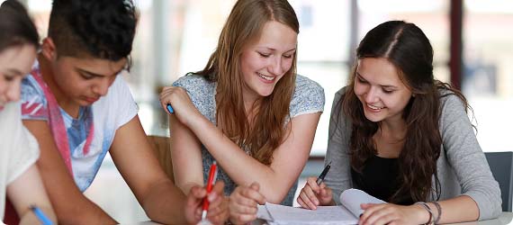 Four students sat around a table working in pairs, looking down whilst sharing textbooks and smiling