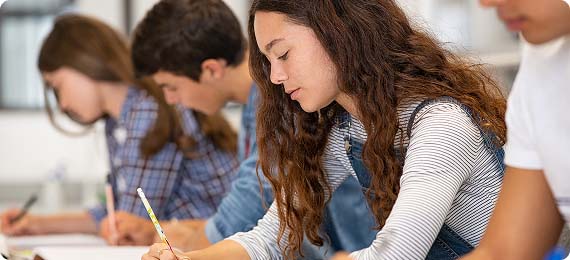Students sat at desks in a row in a classroom with heads down writing in textbooks