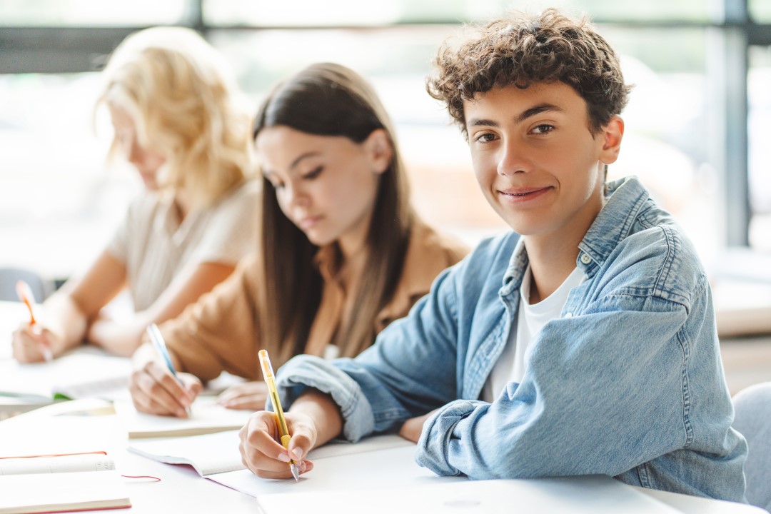 Three students sat in a brightly lit classroom all taking notes with the closest student smiling at the camera