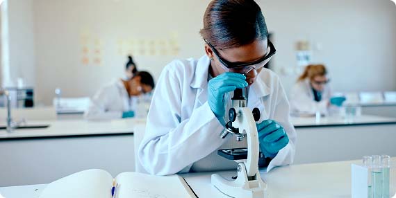 Student in a science lab wearing a lab coat, goggles and gloves looking through a microscope
