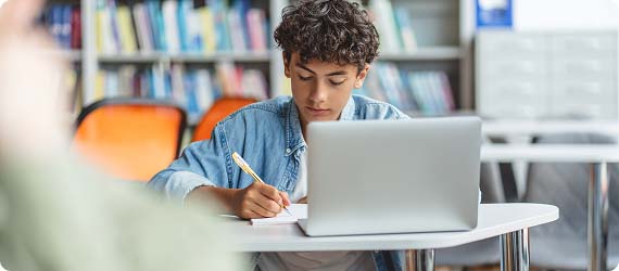 Student sat at a table in library with laptop writing notes