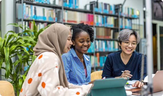 Three students sat at a table in library smiling whilst looking down at a laptop screen together
