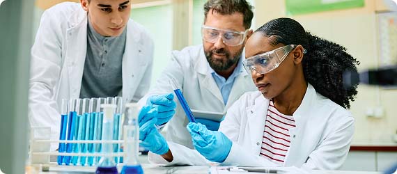 Two students and staff wearing lab coats, goggles and gloves looking at test tubes with blue liquid inside