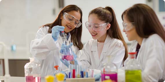 Three students wearing lab coats, goggles and gloves transferring coloured liquid into test tubes