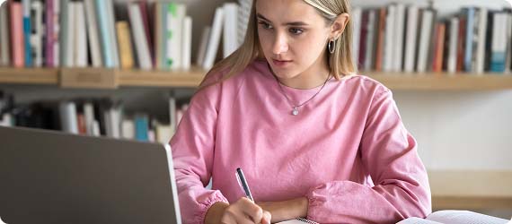 Student sat in library writing in notebook whilst looking at laptop screen