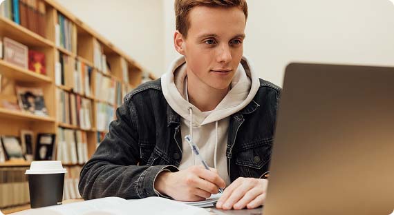 Student sat in library in front of laptop taking notes whilst having a coffee