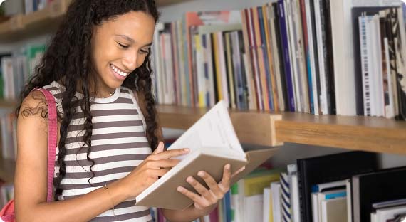 Student stood in library next to shelves of books, smiling whilst looking down and turning the page of a book