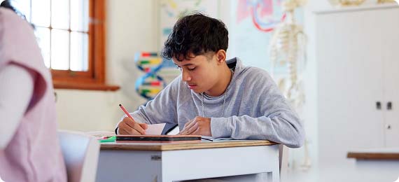 Student sat at a desk writing notes in classroom with skeleton model and atom structure in background