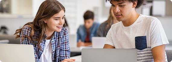 Students sat in a classroom in front of laptops working in pairs discussing work on a screen