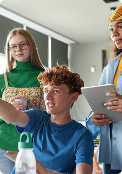 Student wearing blue t-shirt sitting down pointing to the front with two students standing behind him