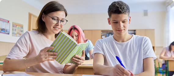 Teacher and students sat in classroom at desks reading from a book and making notes with a pen
