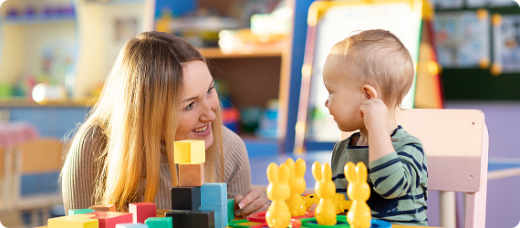 Early years educator sat talking and smiling to a toddler who is sat at a child's table playing with building blocks and toy bunnies