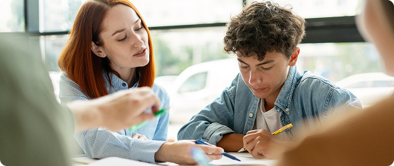 Teacher and student looking at textbook and notes on a desk, whilst the teacher is pointing something out on the notes with a pen