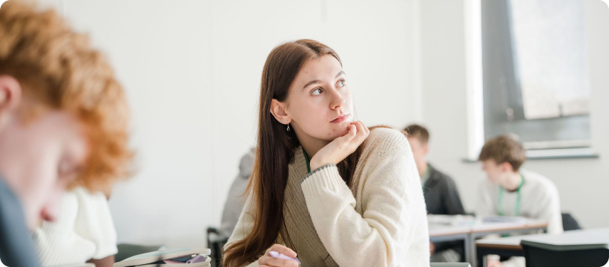 Student wearing white top sitting in the classroom looking towards left with other students at the background