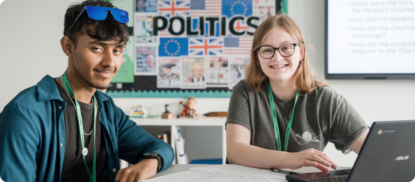 Students sitting in front of laptop smiling with politics news articles wall board at the back