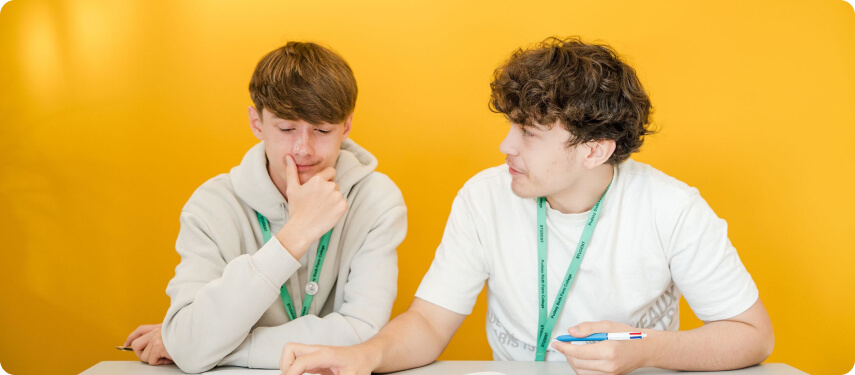 Students wearing green lanyard sitting down having conversation with yellow wall at the back