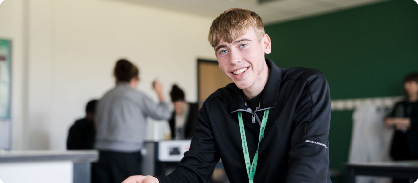 Student in black top wearing green lanyard smiling in the classroom with other students at the background