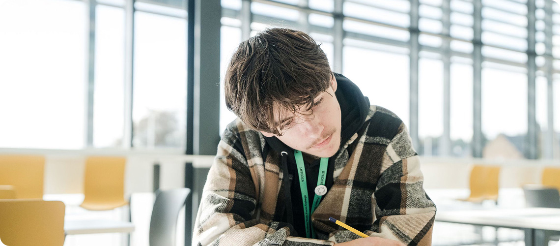Student wearing checked top sitting down holding pen concentrating on notes