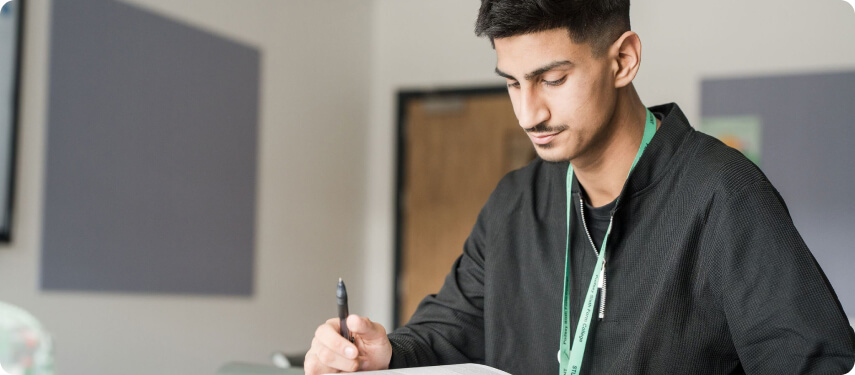 Student wearing green lanyard sitting in classroom holding a pen and reading