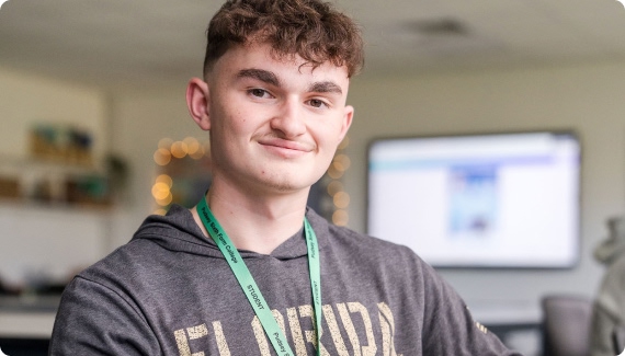 Student in grey top wearing green lanyard smiling with a big screen at the background