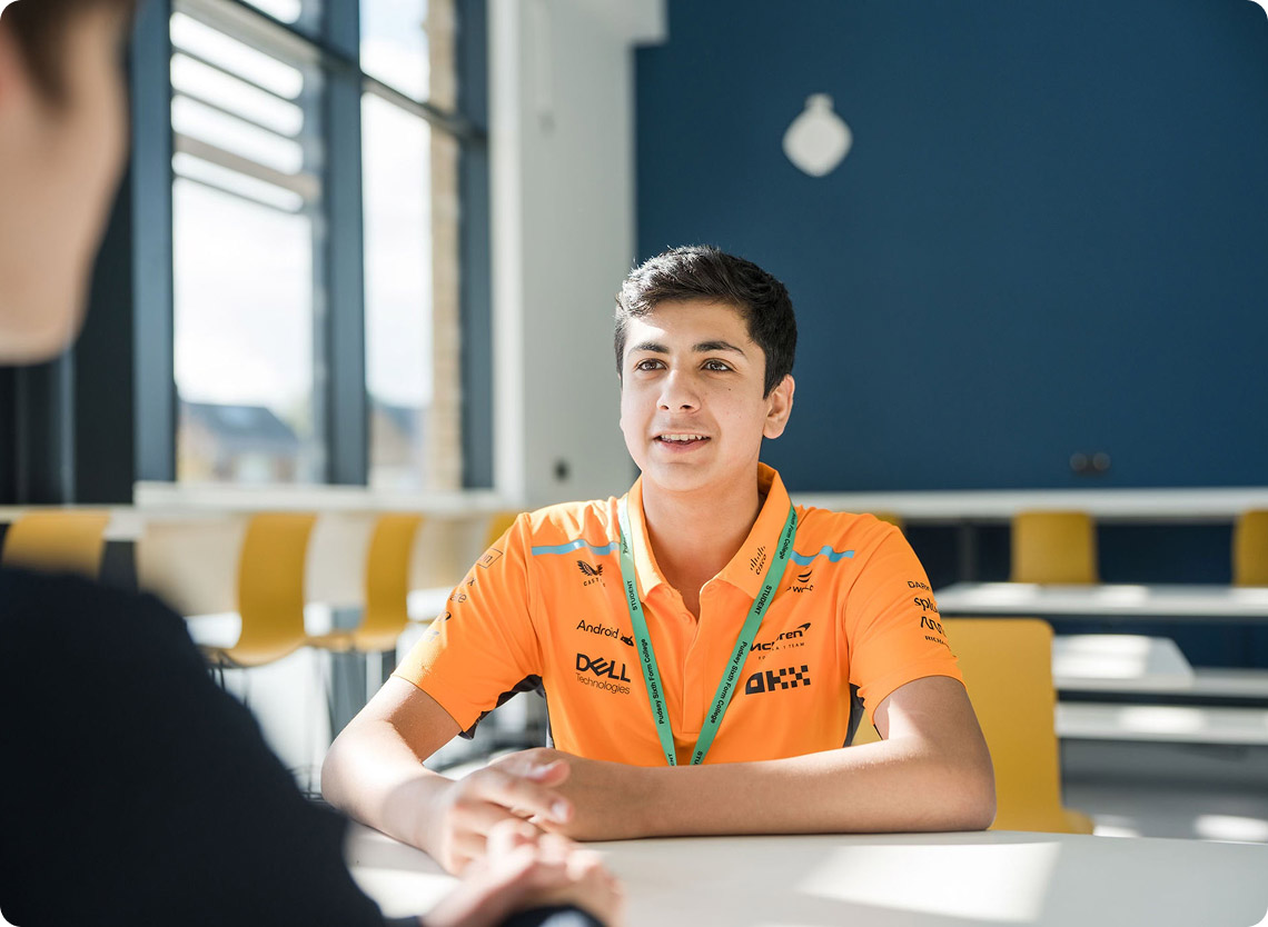 Student wearing orange top green lanyard sitting down talking with another student