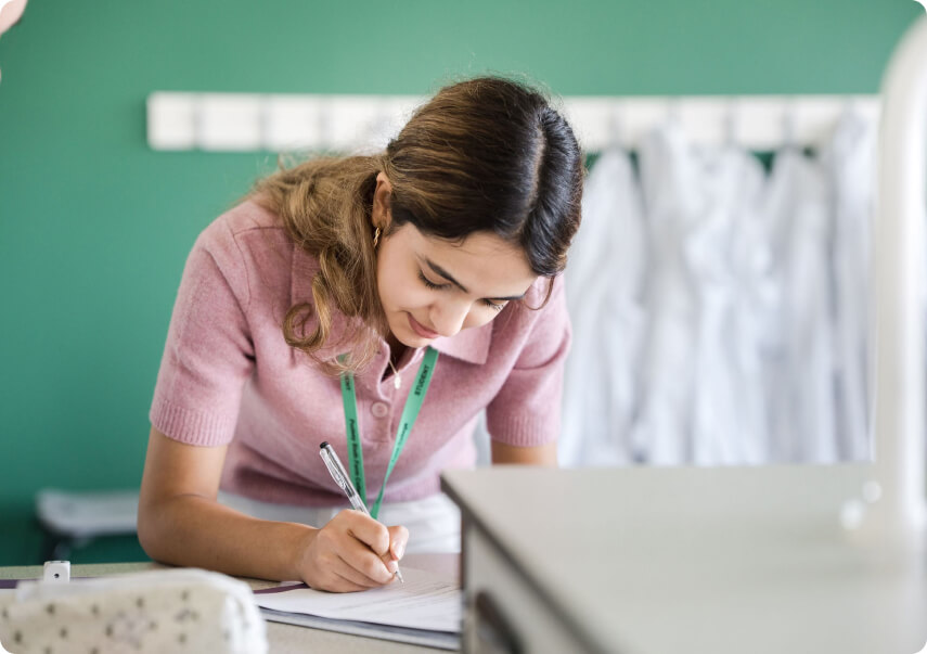 Student in pink top wearing green lanyard bending and making notes in the classroom with some white lab coats hanging on the wall at the background