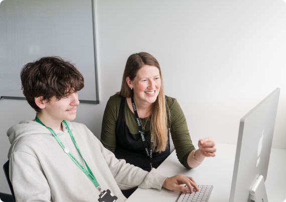 Tutor and student sitting down in classroom looking at computer monitor smiling