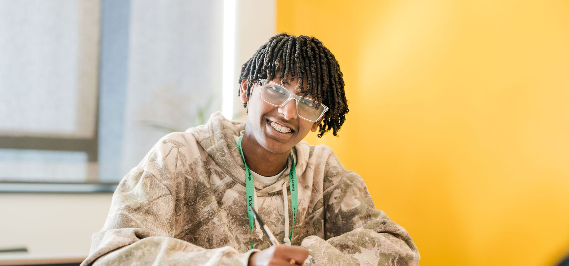 Student sat at a desk in classroom with yellow background, looking up and smiling