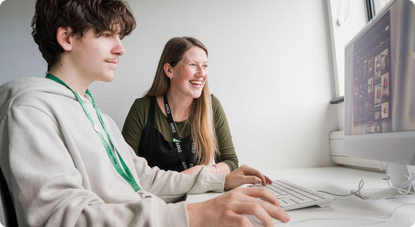 Tutor and student sitting down in classroom looking at computer monitor smiling
