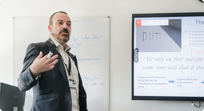 Tutor standing in front of classroom talking with monitor screen and white board at the background