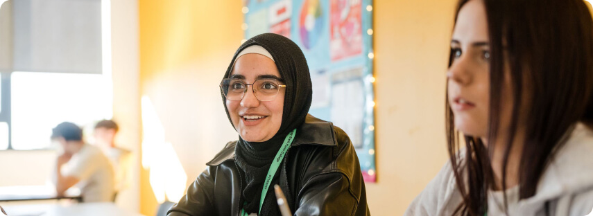 Student wearing hijab headscarf sitting down smiling with other students at the background in the classroom