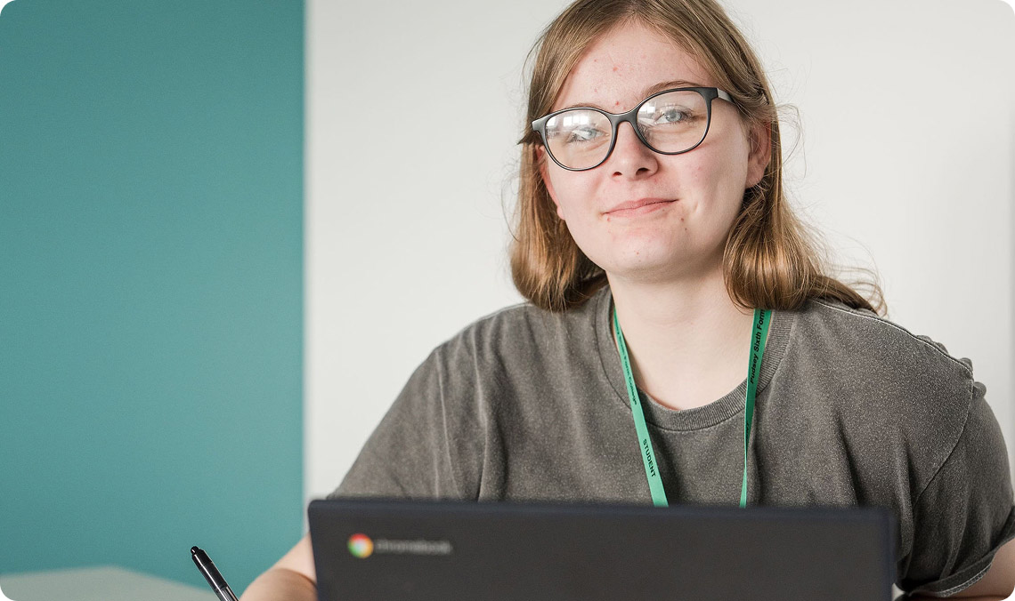 Student wearing grey top and glass in classroom with laptop smiling